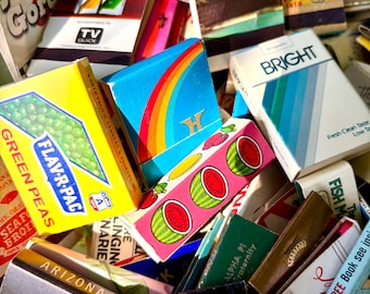 May include: A collection of vintage matchbooks and boxes, featuring a variety of colorful designs and text. Notable items include a box of "Green Peas", a "Bright" cigarette pack, and matchbooks advertising "Sahara" and "The Mint" hotels.