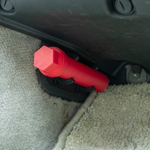 May include: A red, cylindrical car safety device with a hexagonal cap. The device is tucked into the corner of a car's interior.