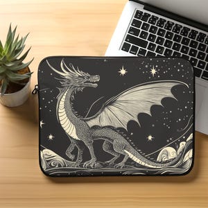 May include: Black laptop sleeve featuring a detailed illustration of a dragon with large wings, set against a starry night sky. The dragon is in shades of gray and cream, with intricate scales and a majestic pose. The sleeve is designed to protect a laptop.