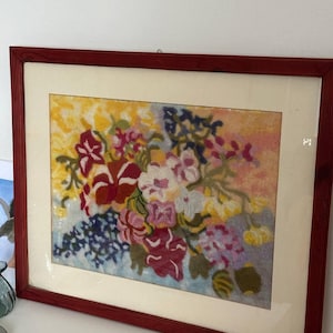 May include: A framed floral needlepoint artwork. The artwork features a bouquet of flowers in shades of red, pink, white, yellow, and blue. The frame is a dark reddish-brown color. The background is a mix of yellow, pink, and blue.