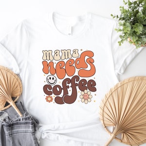 May include: White t-shirt with a retro-style graphic that says "mama needs coffee" in orange, brown, and white. The graphic includes a smiley face and flowers.