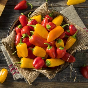 May include: A bowl overflowing with a colorful assortment of fresh, raw mini bell peppers. The peppers are a mix of red, yellow, and orange, with green stems. The bowl sits on a rustic wooden surface, with a burlap liner.