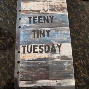 May include: A clear plastic page protector with a woodgrain background. The words "TEENY TINY TUESDAY" are printed in black on the page protector.