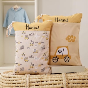 Könnte beinhalten: Zwei beige-gelbe Stoffbeutel mit dem Aufdruck "Hannis". Eine Tasche hat ein Muster aus gelben Fahrzeugen auf beigem Hintergrund. Die andere Tasche hat eine gelbe Autografik und eine gelbe Wolkengrafik.