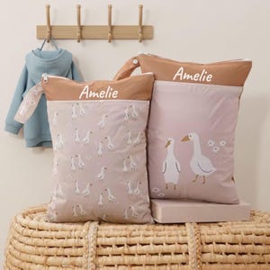 Könnte beinhalten: Zwei personalisierte Baby-Taschen mit dem Aufdruck "Amelie". Eine Tasche zeigt ein Gänsemuster, die andere zwei Gänse mit Blumen. Die Taschen haben einen braunen oberen Teil und einen Reißverschluss. Im Hintergrund hängt ein blauer Kapuzenpullover.