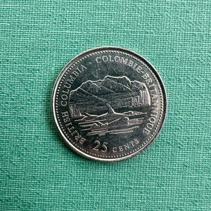 May include: A silver 25-cent coin with a mountain range and a whale design. The coin has the text "BRITISH COLUMBIA" and "COLOMBIE-BRITANNIQUE". The coin is on a green textured surface.