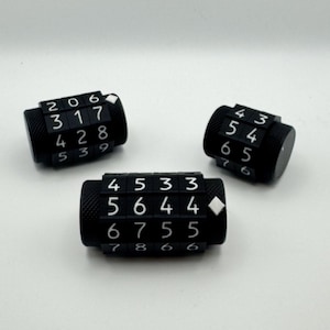 May include: Three black combination lock dials with white numbers are arranged on a white background. Each dial has a series of numbers visible, with a diamond-shaped indicator on one. The dials are cylindrical.