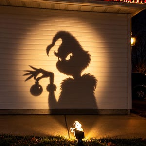 Pode incluir: Um projetor de luz no chão projeta uma grande sombra escura de um personagem com chapéu pontudo e dedos longos segurando um ornamento redondo no revestimento horizontal branco de uma casa durante a noite.