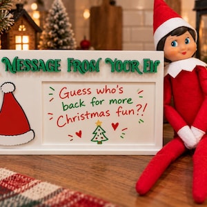 Könnte beinhalten: Ein weißes rechteckiges Schild mit dem grünen Text A MESSAGE FROM YOUR ELF und einem roten Weihnachtsmannmützen-Symbol. In der Mitte steht der Text Guess who's back for more Christmas fun?! mit einem kleinen Baum und Herzen. Eine Elfenpuppe sitzt daneben.