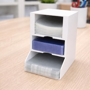 May include: A white, three-tiered desk organizer with stacks of clear and blue plastic sheets. The organizer sits on a light wood-grain surface. The top shelf holds a stack of clear sheets, the middle shelf holds blue sheets, and the bottom shelf holds clear sheets.