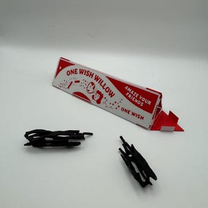 Puede incluir: Una caja triangular de cart&oacute;n blanca y roja etiquetada como ONE WISH WILLOW con el texto AMAZE YOUR FRIENDS y ONE WISH, acompa&ntilde;ada de dos peque&ntilde;os objetos de pl&aacute;stico negro, de forma irregular, huecos y similares a ramas.