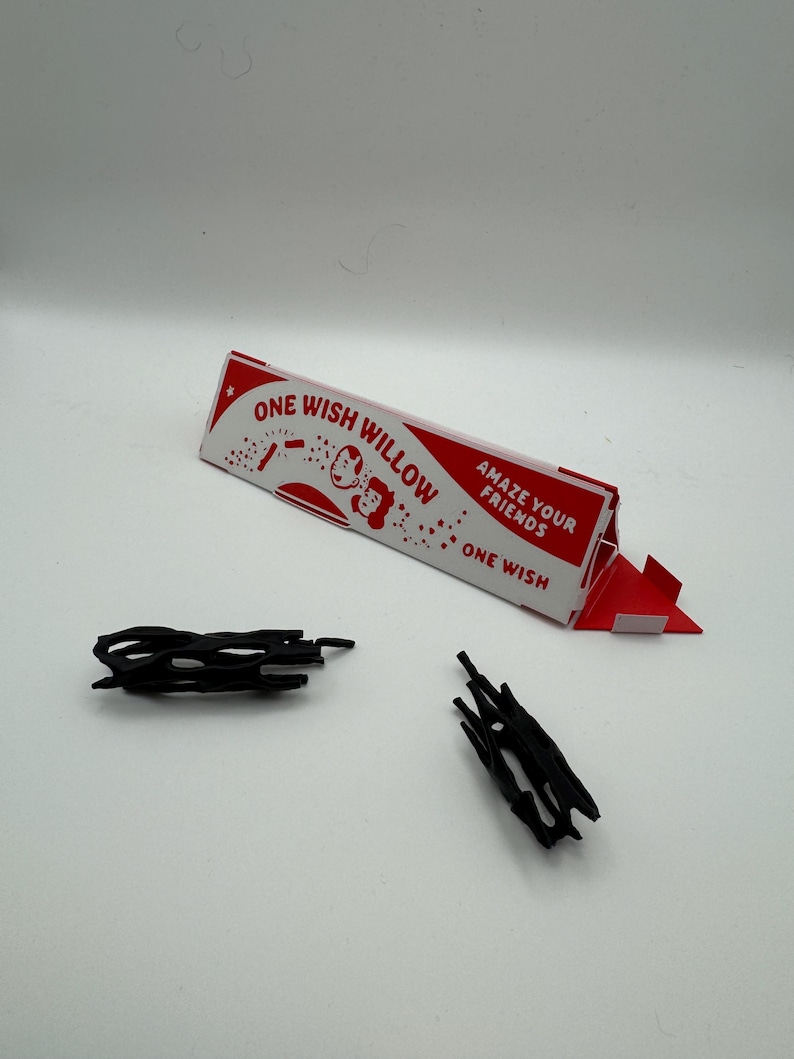 Puede incluir: Una caja triangular de cart&oacute;n blanca y roja etiquetada como ONE WISH WILLOW con el texto AMAZE YOUR FRIENDS y ONE WISH, acompa&ntilde;ada de dos peque&ntilde;os objetos de pl&aacute;stico negro, de forma irregular, huecos y similares a ramas.