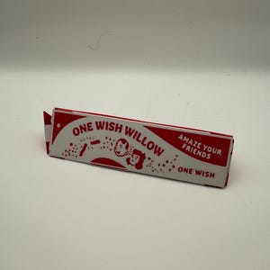 Puede incluir: Una caja rectangular de papel roja y blanca etiquetada como ONE WISH WILLOW con el texto AMAZE YOUR FRIENDS y ONE WISH. La caja presenta una ilustraci&oacute;n gr&aacute;fica de dos rostros sonrientes rodeados de peque&ntilde;as estrellas y confeti.