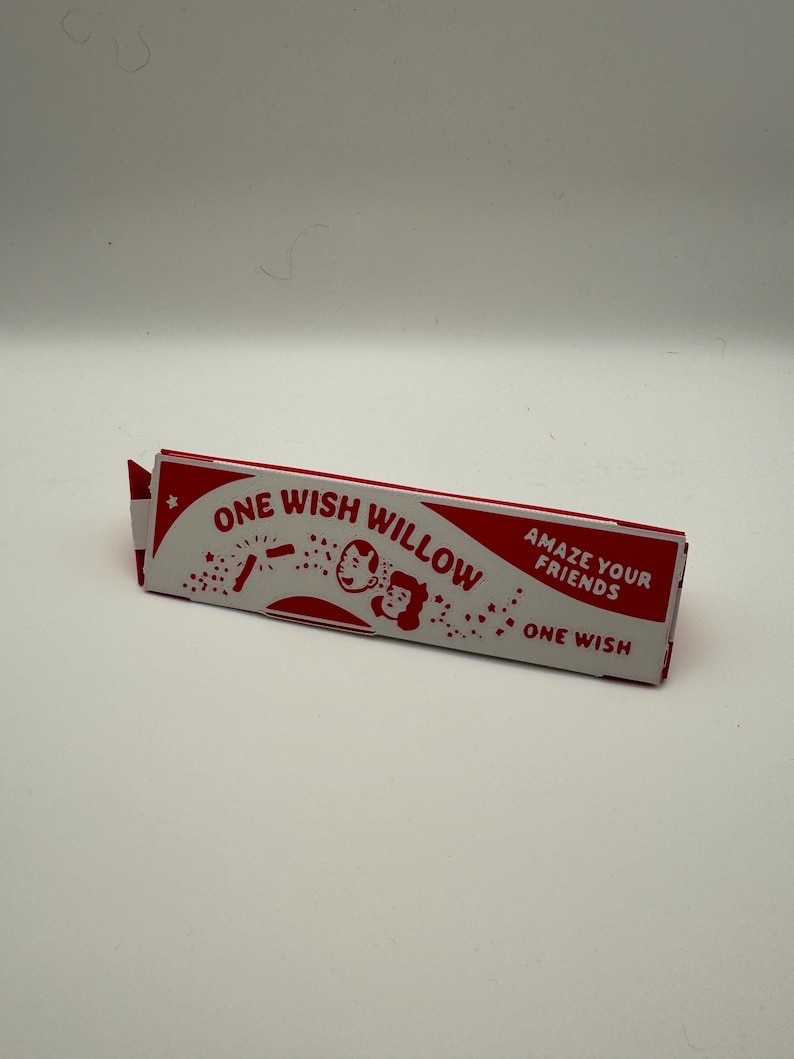 Puede incluir: Una caja rectangular de papel roja y blanca etiquetada como ONE WISH WILLOW con el texto AMAZE YOUR FRIENDS y ONE WISH. La caja presenta una ilustraci&oacute;n gr&aacute;fica de dos rostros sonrientes rodeados de peque&ntilde;as estrellas y confeti.