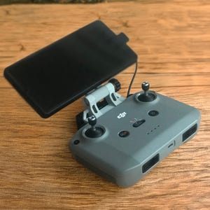 May include: A gray drone remote control with two joysticks, buttons, and a black screen attached. The remote is on a wooden surface. The remote control has the DJI logo.