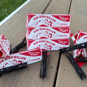 May include: A stack of rectangular white boxes with red accents labeled ONE WISH WILLOW and AMAZE YOUR FRIENDS, accompanied by several black, elongated, lattice-patterned plastic objects scattered around the base of the boxes.