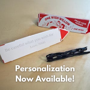 Puede incluir: Una tarjeta de papel blanco con detalles en rojo que presenta el texto ONE WISH WILLOW y AMAZE YOUR FRIENDS, junto a una segunda tarjeta blanca impresa con Be careful what you wish for Love, Nikki, y un peque&ntilde;o objeto cil&iacute;ndrico negro poroso.