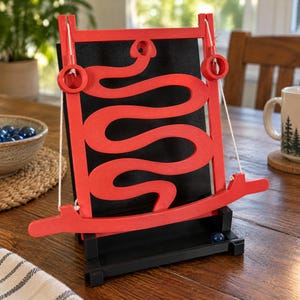 May include: A red wooden labyrinth game featuring a winding path mounted on a black board. The game includes two white strings attached to a horizontal handle, allowing for movement. A single blue marble rests in the black base tray.