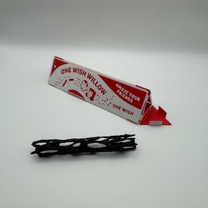Puede incluir: Una caja de presentaci&oacute;n triangular de cart&oacute;n rojo y blanco etiquetada como ONE WISH WILLOW con el texto AMAZE YOUR FRIENDS y ONE WISH, acompa&ntilde;ada de un objeto negro, alargado y cil&iacute;ndrico con una estructura de red hueca.