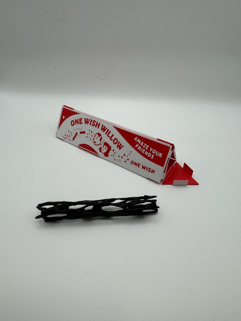 Puede incluir: Una caja de presentaci&oacute;n triangular de cart&oacute;n rojo y blanco etiquetada como ONE WISH WILLOW con el texto AMAZE YOUR FRIENDS y ONE WISH, acompa&ntilde;ada de un objeto negro, alargado y cil&iacute;ndrico con una estructura de red hueca.