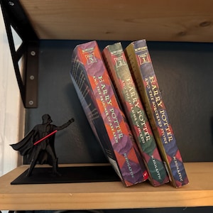 May include: Three Harry Potter books with colorful covers, leaning against a black Darth Vader figurine with a red lightsaber. The books are titled "Harry Potter and the Sorcerer's Stone", "Harry Potter and the Chamber of Secrets", and "Harry Potter and the Prisoner of Azkaban".