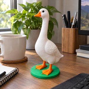 May include: A white goose figurine with an orange beak and orange webbed feet standing on a circular green base. The goose has a smooth white body, a long neck, and a small black eye. The figurine is positioned upright on a flat surface.