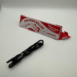 Puede incluir: Un objeto negro de forma alargada con estructura de celos&iacute;a y centro hueco, situado junto a una caja triangular de cart&oacute;n roja y blanca con la inscripci&oacute;n ONE WISH WILLOW y el texto AMAZE YOUR FRIENDS y ONE WISH en la parte frontal.