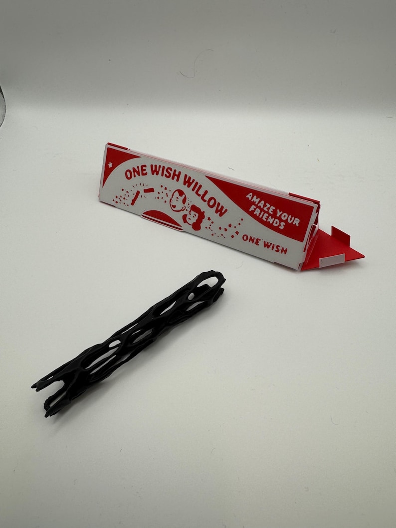 Puede incluir: Un objeto negro de forma alargada con estructura de celos&iacute;a y centro hueco, situado junto a una caja triangular de cart&oacute;n roja y blanca con la inscripci&oacute;n ONE WISH WILLOW y el texto AMAZE YOUR FRIENDS y ONE WISH en la parte frontal.