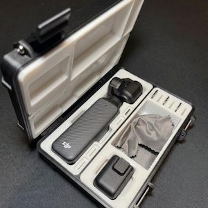 May include: Black and white hard case with a DJI camera and accessories. The case is open, revealing a camera, a charging base, a cleaning cloth, and other small items. The case has a white interior and a black exterior.
