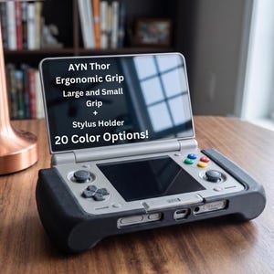Puede incluir: Consola de juegos portátil con un agarre ergonómico negro y el texto "AYN Thor Ergonomic Grip Large and Small Grip + Stylus Holder 20 Color Options!". La consola tiene un cuerpo plateado con botones de colores.