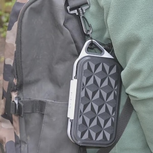 May include: A black and gray geometric patterned portable speaker is attached to a black backpack with a camouflage design. The speaker is hanging from a black strap and has a white accent on the side.