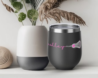 May include: A matte charcoal gray stainless steel tumbler with a clear lid. The tumbler features the name ashley in pink cursive script followed by a white and pink lacrosse stick graphic with a pink ball inside the mesh pocket.