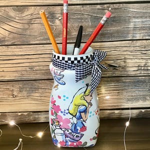 May include: A white ceramic vase with a black and white checkered ribbon around the top. The vase is decorated with a scene from Alice in Wonderland, featuring Alice, a teacup, and the words "Drink Me". There are four pencils in the vase, two red, one yellow, and one black.