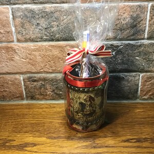 Puede incluir: Un porta lápices rojo y dorado con un diseño de Alicia en el País de las Maravillas. El porta lápices está envuelto en una cinta roja y blanca y celofán transparente.