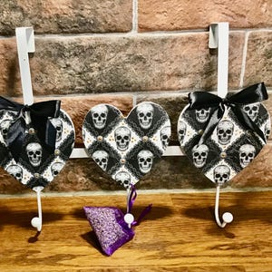 Peut inclure: Trois crochets en forme de cœur blancs avec des motifs de crânes noirs et blancs et des nœuds noirs. Les crochets sont fixés à une barre en métal blanc. Un petit sachet violet est suspendu à l'un des crochets.