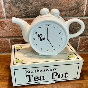Puede incluir: Una tetera de cerámica blanca con forma de reloj, con asa, pico y dos pequeños pomos en la parte superior. La esfera muestra números negros, manecillas marcando las cinco y el texto "TEA TIME". Se apoya sobre una caja con la etiqueta "Earthenware Tea Pot".