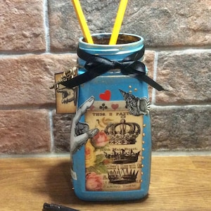 Puede incluir: Un tarro de vidrio azul con una cinta negra y detalles dorados. El tarro está decorado con recortes de papel de una corona, flores y una mano. El tarro está lleno de dos lápices amarillos y tiene el texto "THOS. E. PAXTON" en él.