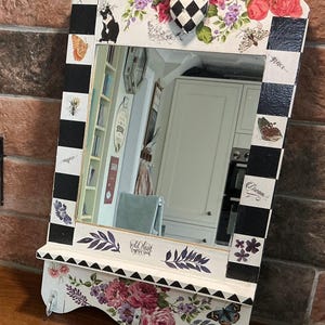 May include: A decorative wall mirror with a shelf and hooks. The mirror frame is white with black and white checkered accents. It features floral and butterfly decals, with the words "fearless", "inspire", "grace", "dream", and "Wild Heart Gypsy Soul".