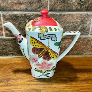 Könnte beinhalten: Eine weiße Keramikteekanne mit quadratischem Körper, rotem Deckel, schwarz-weiß kariertem Henkel und Ausguss. Die Oberfläche ist mit bunten Blumenmustern, grünen Ranken und einem großen gelben Nachtfalter verziert, der den Schriftzug STUNNING auf einem Banner trägt.