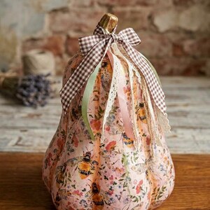 May include: A decorative gourd featuring a light pink surface covered in a floral and bee pattern. The top is adorned with a brown and white chequered ribbon, a light green ribbon, a pink ribbon, and a white lace trim tied around the stem.