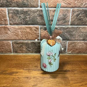 May include: A light blue decorative reed diffuser with floral designs and a decorative bow. The diffuser has light blue reeds and is placed on a wooden surface in front of a brick wall.