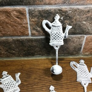 May include: Set of three white cast iron teapot hooks. Each hook features a detailed teapot design with a distressed finish. The hooks are perfect for hanging towels, robes, or other items in a kitchen or bathroom.