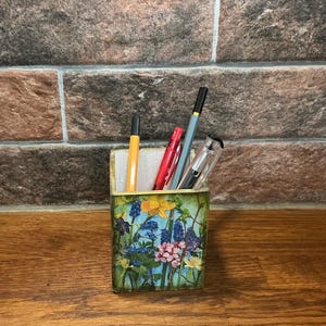Może przedstawiać: Drewniany organizer na ołówki z kwiatowym wzorem z niebieskimi, różowymi, żółtymi i zielonymi kwiatami. Organizer zawiera cztery długopisy, w tym jeden czerwony, jeden czarny i dwa szare.