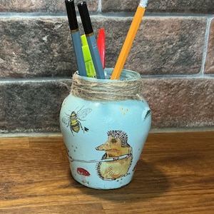 Puede incluir: Un porta lápices de cerámica azul con un diseño pintado de un erizo, una abeja y setas. El porta lápices está envuelto con cuerda y tiene tres lápices dentro.