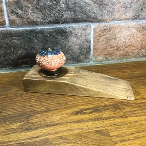 May include: A wooden doorstop with a decorative ceramic knob. The knob is peach-colored with white and blue floral designs.