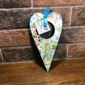May include: A wooden heart-shaped ornament with a blue ribbon. The ornament is decorated with a floral pattern and a picture of Peter Rabbit. The text "Admit One" is visible on the ornament.