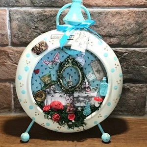 Puede incluir: Un reloj blanco y azul con una cara redonda y un lazo azul en la parte superior. La cara del reloj tiene un fondo blanco con lunares azules y una escena de Alicia en el País de las Maravillas. La escena incluye al Sombrerero Loco, la Liebre de Marzo, el Lirón y el Gato de Cheshire. El reloj tiene un soporte de metal azul.