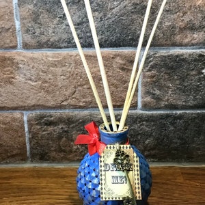 Puede incluir: Un difusor azul texturizado con varillas de caña. La botella tiene una etiqueta que dice "DRINK ME!" y un dije de llave. Un lazo rojo adorna el cuello de la botella. El fondo es una pared de ladrillos.