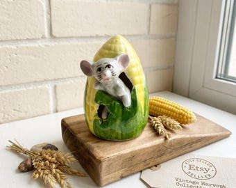 Op de afbeelding: Een keramieken beeldje van een grijze muis die uit een gele maïskolf met groene bladeren kijkt. De muis heeft roze oren en zwarte ogen. De tekst op het label luidt Etsy Seller Vintage Harvest Collection #3.