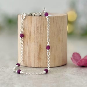 May include: A delicate silver chain bracelet featuring small, round, deep magenta beads spaced evenly along the links. The bracelet includes a secure lobster claw clasp and is constructed with polished silver metal links that catch the light.
